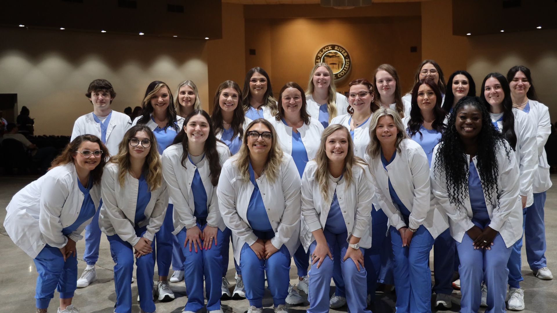 A large group photo of Practical Nursing Program graduates in matching blue scrubs and white jackets, posing together in front of the Black River Technical College seal.